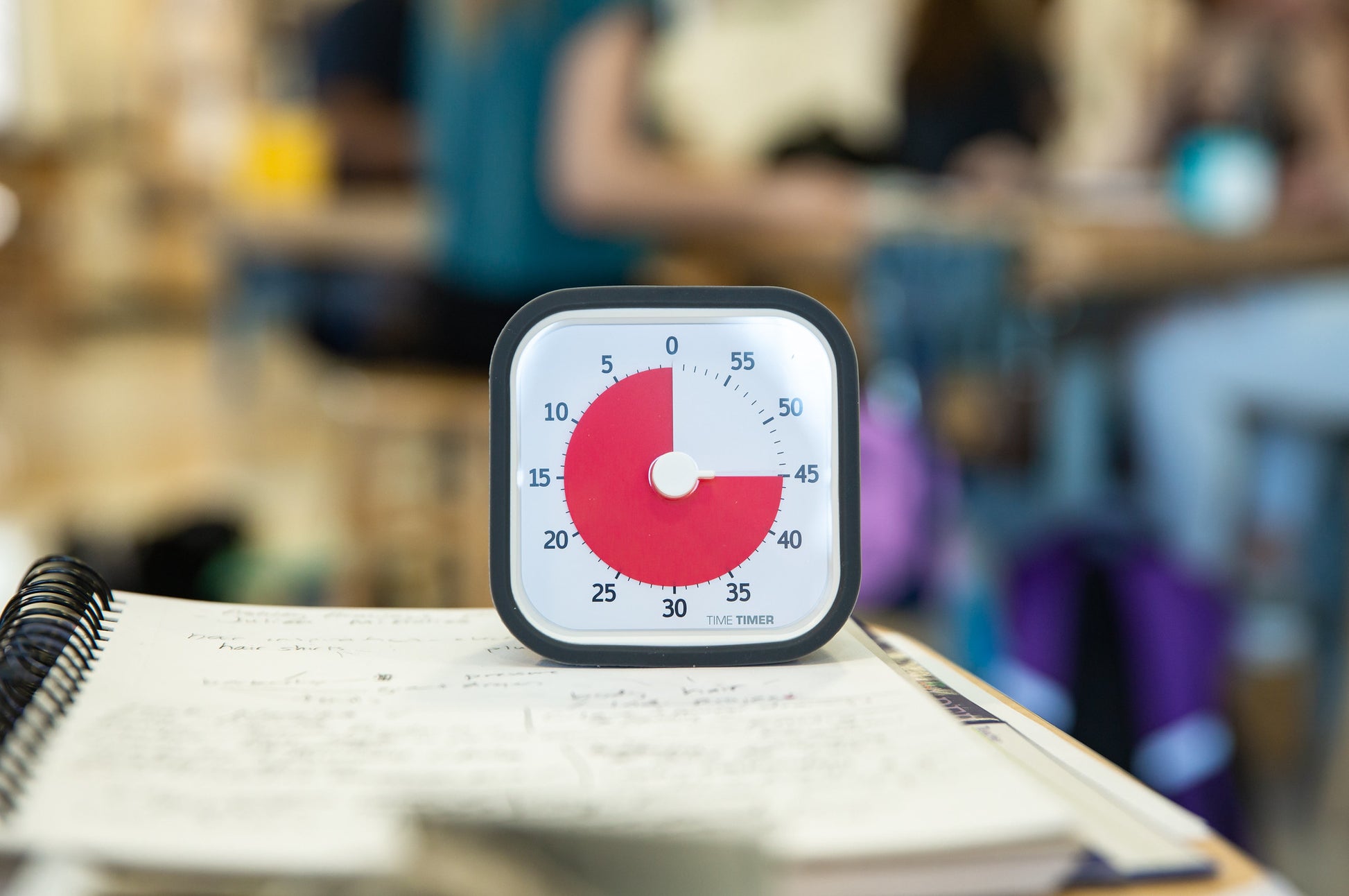 A Time Timer MOD [Charcoal] by Time Timer, showing 35 minutes left with its red countdown, sits on a notebook filled with handwritten notes. In the blurred background, people are seated at tables in a study environment.