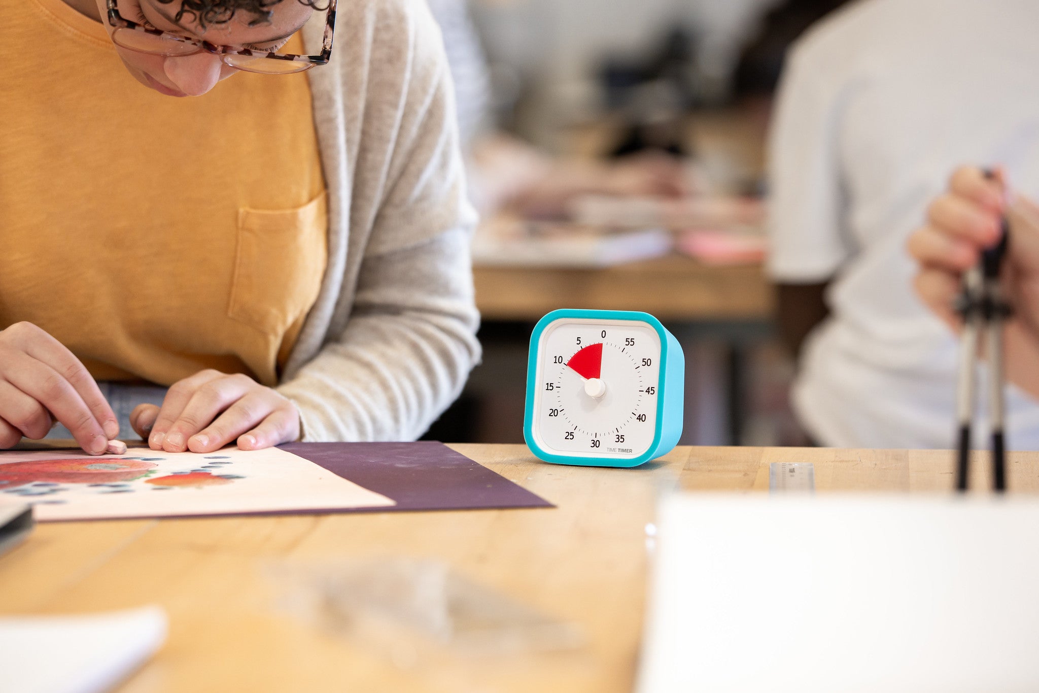 A student works on a project at a desk, with a Time Timer MOD [Sky Blue] (60 min) by Time Timer—set to 17 minutes and in its silicone case—on the table. Another person in the background also uses this time management tool.