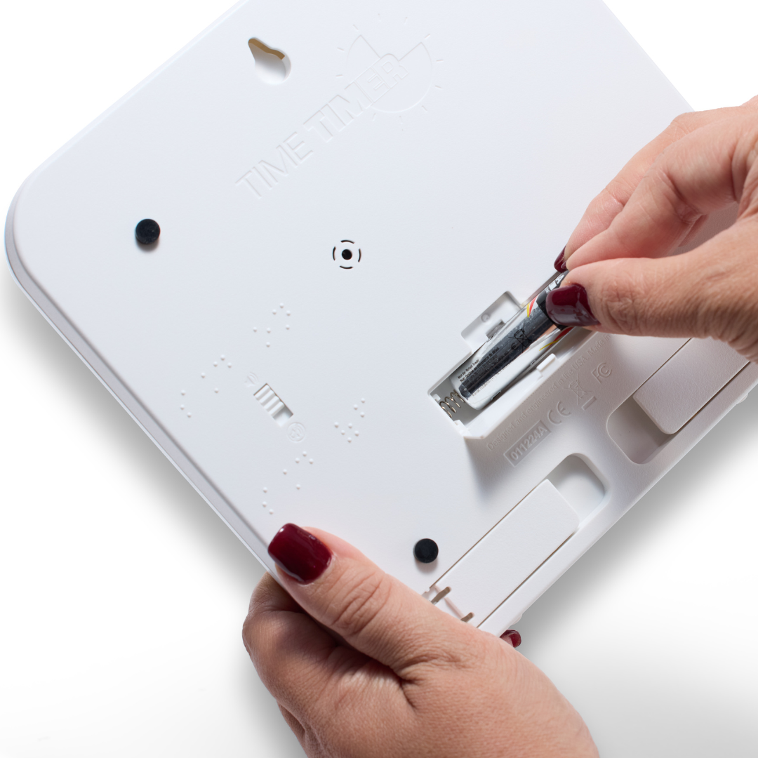 A person with red nail polish inserts an AA battery into the back of a Time Timer Braille (8 Inch) [Red Disk] by Time Timer. The open compartment shows braille markings, highlighting its accessible tactile timer design.