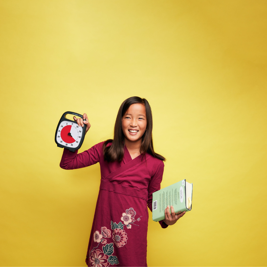 A smiling girl in a maroon dress holds a Time Timer Plus (60 min) by Time Timer in one hand and a book in the other, standing before a bright yellow backdrop—a playful way to show focus tools for effective time management.