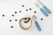 A bowl of yogurt with blueberries sits beside scattered berries and a KiddiKutter Kids Cutlery Set [Sky Blue] by KiddiKutter, featuring blue-handled self-feeding utensils.