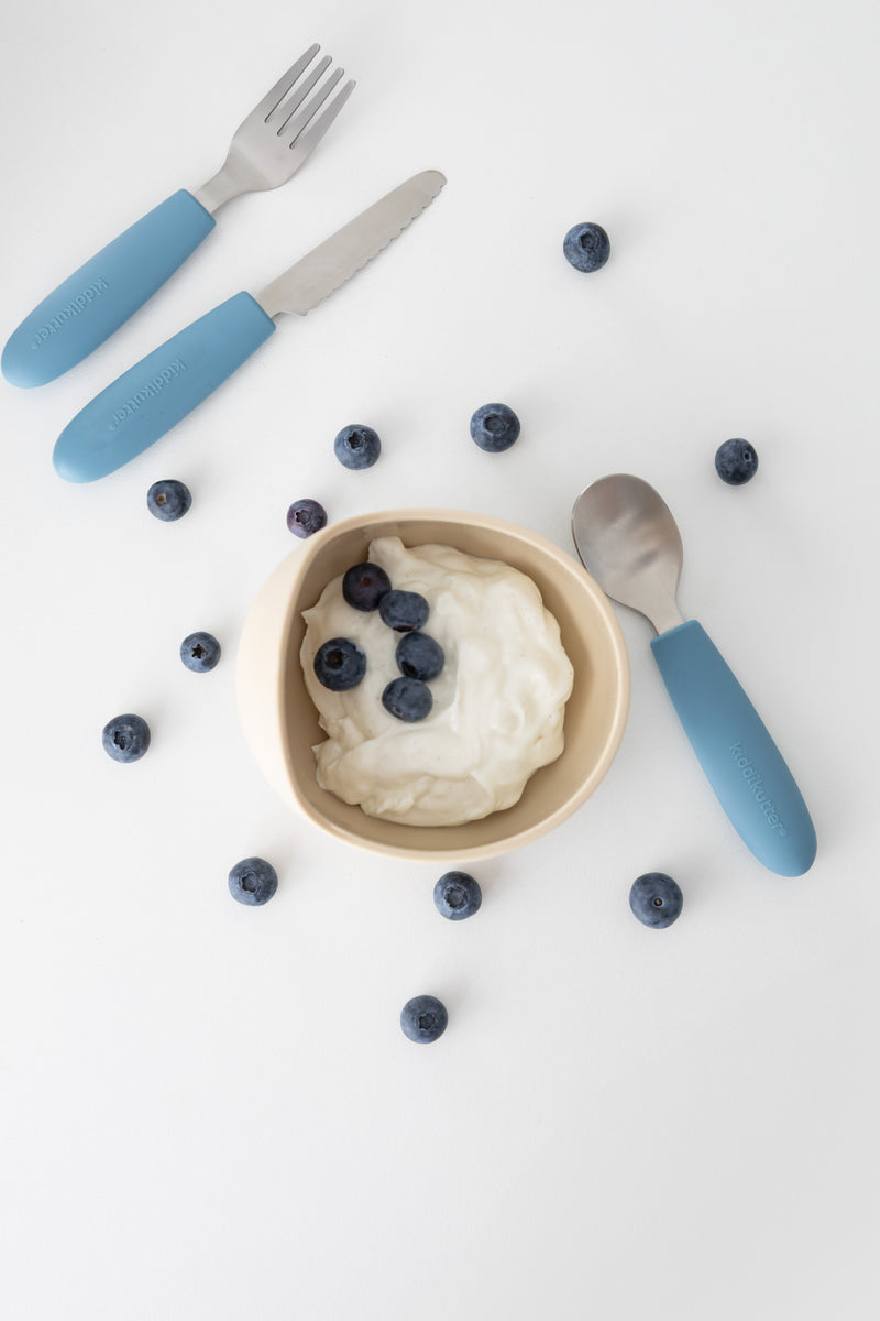 A bowl of yogurt with blueberries sits on a white surface surrounded by scattered berries, while the KiddiKutter Kids Cutlery Set [Peach Fuzz] by KiddiKutter—a fork, knife, and spoon—completes the scene.