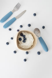 A small bowl of yogurt with blueberries is surrounded by scattered berries. Nearby, a KiddiKutter Kids Cutlery Set [Sky Blue] by KiddiKutter, with blue silicone handles, rests on a white surface.