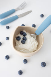 A beige bowl of yogurt with blueberries sits on a white surface, surrounded by berries. In the background is the KiddiKutter Kids Cutlery Set [Sky Blue] by KiddiKutter, featuring self-feeding utensils with blue handles.