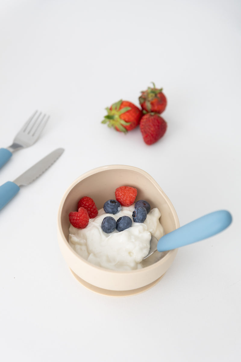A beige bowl of yogurt with blueberries and raspberries, a blue spoon, and fresh strawberries sits on a white surface with a KiddiKutter Kids Cutlery Set [Sky Blue] by KiddiKutter in the background.