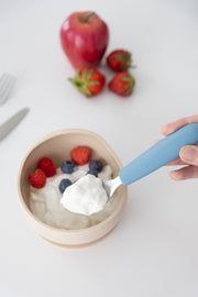 A hand holds the KiddiKutter Kids Cutlery Set [Sky Blue] spoon above a bowl of yogurt topped with raspberries, blueberries, and strawberries. An apple and three strawberries are in the background on a white surface. Brand: KiddiKutter.
