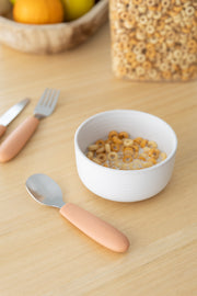 A white bowl of cereal with milk sits on a light wooden table beside the KiddiKutter Kids Cutlery Set [Peach Fuzz]. In the background are a cereal container and a wooden fruit bowl. Brand: KiddiKutter.
