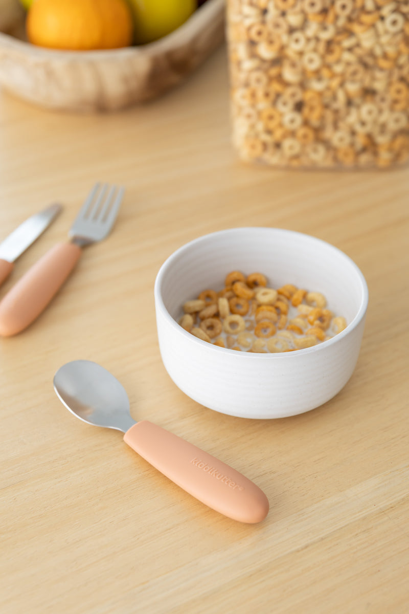 A white bowl of cereal with milk sits on a light wooden table beside the KiddiKutter Kids Cutlery Set [Peach Fuzz]. In the background are a cereal container and a wooden fruit bowl. Brand: KiddiKutter.