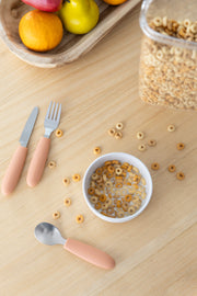 A small white bowl of cereal with milk sits on a wooden table, surrounded by scattered cereal pieces. Nearby, a KiddiKutter Kids Cutlery Set [Peach Fuzz] by KiddiKutter and a tray of fruit complete the scene.