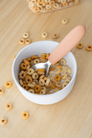 A white bowl of milk with multigrain O-shaped cereal sits on a light wooden surface. A KiddiKutter Kids Cutlery Set [Peach Fuzz] spoon by KiddiKutter rests inside, while some cereal pieces are scattered on the table.
