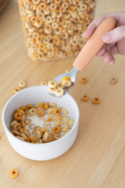 A hand holds a spoon from the KiddiKutter Kids Cutlery Set [Peach Fuzz] over a bowl of cereal and milk, with a cereal container and scattered pieces on a wooden table. Brand: KiddiKutter.
