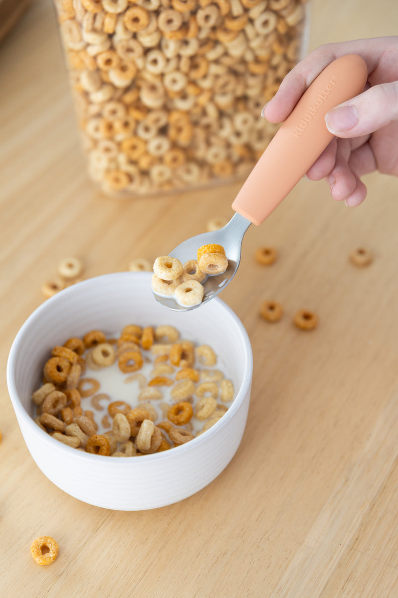 A hand holds a spoon from the KiddiKutter Kids Cutlery Set [Peach Fuzz] over a bowl of cereal and milk, with a cereal container and scattered pieces on a wooden table. Brand: KiddiKutter.