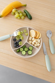 A divided plate with sliced cucumber, blueberries, green grapes, cheese cubes, and round crackers sits on a wooden table. Nearby is the KiddiKutter Kids Cutlery Set [Sage] by KiddiKutter, plus extra grapes, a banana, and a cucumber.