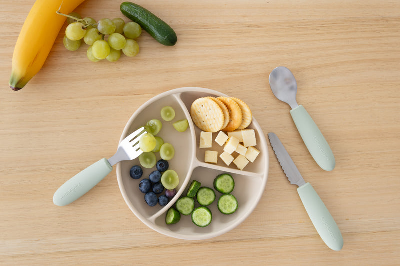 A divided plate with crackers, cheese cubes, cucumber slices, grapes, and blueberries sits on a wooden table. Nearby are a banana, green grapes, a cucumber, and the KiddiKutter Kids Cutlery Set [Sage] by KiddiKutter—ideal for little hands learning to eat.