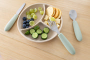A beige divided plate holds sliced grapes, blueberries, cucumber, cheese cubes, and round crackers. The KiddiKutter Kids Cutlery Set [Sage] by KiddiKutter rests nearby—perfect for encouraging self-feeding.