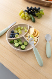 A beige divided plate with cucumber slices, blueberries, green grapes, cheese cubes, and crackers sits on a wooden table next to a KiddiKutter Kids Cutlery Set [Sage] by KiddiKutter; extra grapes are visible in the background.