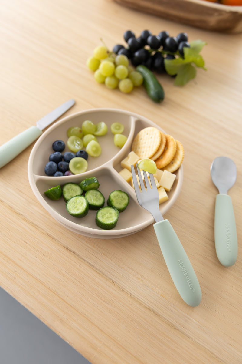 A beige divided plate with cucumber slices, blueberries, green grapes, cheese cubes, and crackers sits on a wooden table next to a KiddiKutter Kids Cutlery Set [Sage] by KiddiKutter; extra grapes are visible in the background.