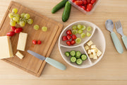A wooden table holds cheese, grapes, tomatoes, a knife, and a tray with sliced cucumbers, cheese cubes, grapes, and tomatoes—perfect for little hands using the KiddiKutter Kids Cutlery Set [Sage] by KiddiKutter.