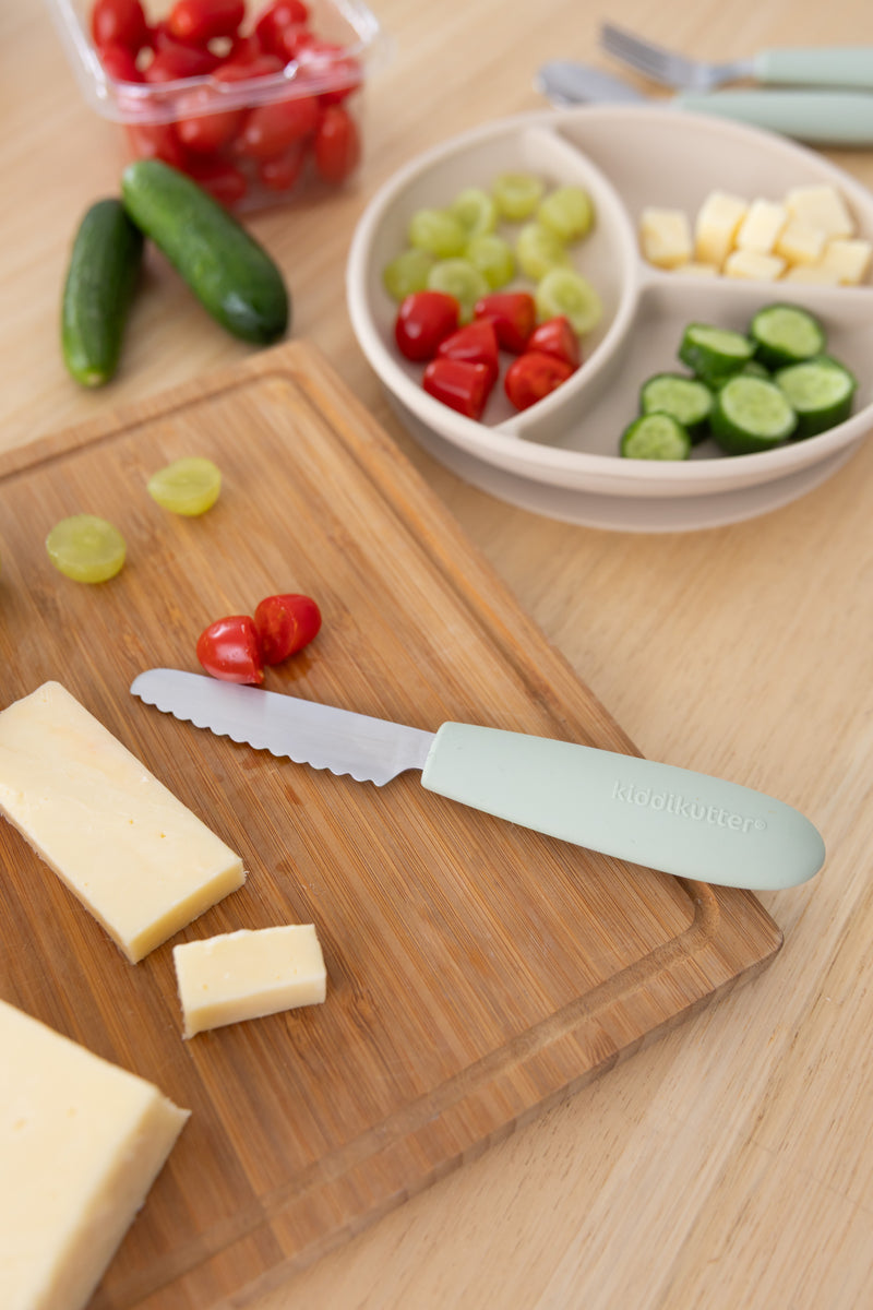 A KiddiKutter Kids Cutlery Set [Sage] knife by KiddiKutter sits on a wooden board with sliced cheese, grape tomatoes, and grapes. A divided plate holds cheese cubes, tomatoes, grapes, and cucumber slices. Mini cucumbers are in the background.