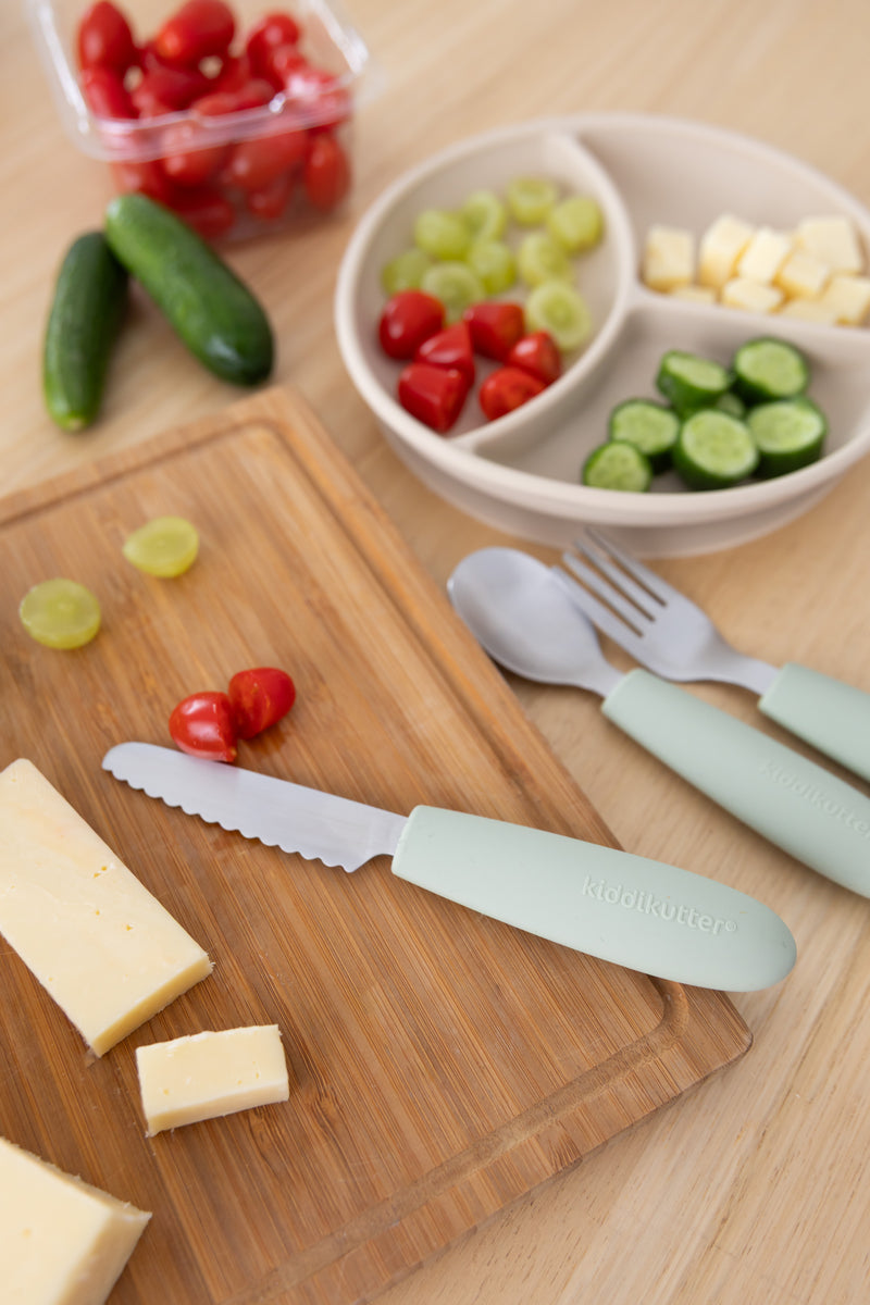 A wooden cutting board holds sliced cheese, cherry tomatoes, and grapes. Nearby is the KiddiKutter Kids Cutlery Set [Sage] by KiddiKutter and a plate with grapes, cheese cubes, cucumbers, tomatoes, and self-feeding utensils.