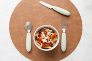 A bowl of pasta with tomato sauce and cheese sits on a brown placemat. Next to it, the KiddiKutter Kids Cutlery Set [Sage] by KiddiKutter, featuring charming light green handles, includes a fork, spoon, and serrated knife.