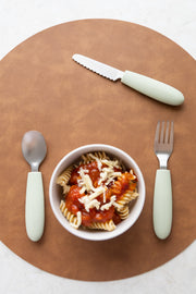 A bowl of pasta with tomato sauce and cheese sits on a round brown placemat, with the KiddiKutter Kids Cutlery Set [Sage] by KiddiKutter—ideal self-feeding utensils for children—arranged around it.