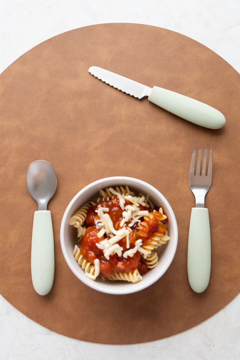 A bowl of pasta with tomato sauce and cheese sits on a round brown placemat, with the KiddiKutter Kids Cutlery Set [Sage] by KiddiKutter—ideal self-feeding utensils for children—arranged around it.