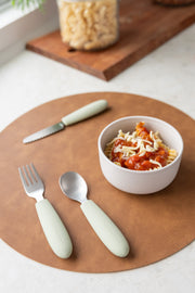 A bowl of pasta with tomato sauce and cheese sits on a brown placemat beside a fork, spoon, and knife from the KiddiKutter Kids Cutlery Set [Sage] by KiddiKutter. A jar of uncooked pasta is visible in the background.
