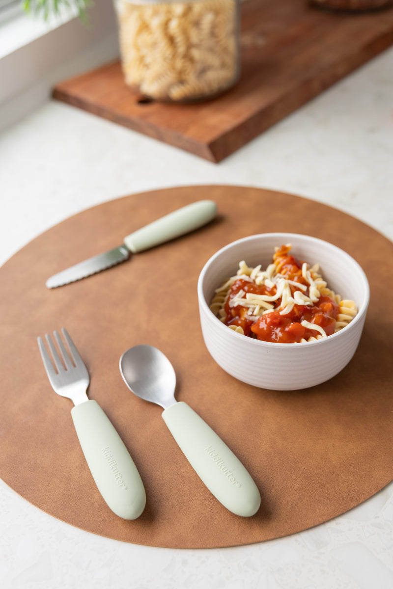 A bowl of pasta with tomato sauce and cheese sits on a brown placemat beside a fork, spoon, and knife from the KiddiKutter Kids Cutlery Set [Sage] by KiddiKutter. A jar of uncooked pasta is visible in the background.