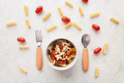 A bowl of spiral pasta with tomato sauce and cheese sits between a fork and spoon from the KiddiKutter Kids Cutlery Set [Peach Fuzz] by KiddiKutter, with uncooked pasta spirals and cherry tomatoes scattered on a light surface nearby.