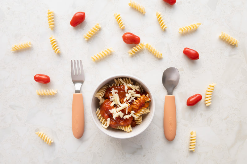 A bowl of spiral pasta with tomato sauce and cheese sits between a fork and spoon from the KiddiKutter Kids Cutlery Set [Peach Fuzz] by KiddiKutter, with uncooked pasta spirals and cherry tomatoes scattered on a light surface nearby.