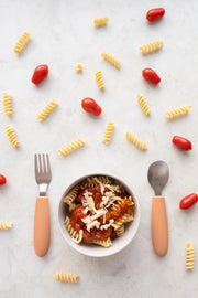 A bowl of fusilli with tomato sauce and cheese sits between a fork and spoon from the KiddiKutter Kids Cutlery Set [Peach Fuzz] by KiddiKutter. Uncooked pasta spirals and grape tomatoes are scattered on the white surface around it.