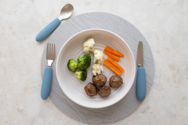 A white plate with three meatballs, steamed broccoli, cauliflower, and carrot sticks sits on a round gray placemat. On either side is the KiddiKutter Kids Cutlery Set [Sky Blue] by KiddiKutter.