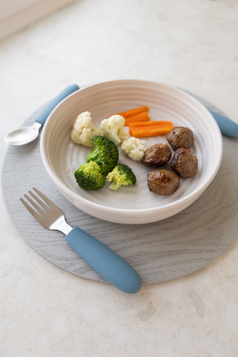 A white bowl of steamed broccoli, cauliflower, carrots, and meatballs sits on a gray placemat. Next to it are a fork and spoon from the KiddiKutter Kids Cutlery Set [Sky Blue] by KiddiKutter.