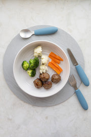 A white bowl with broccoli, cauliflower, carrot sticks, and three meatballs sits on a gray placemat. The KiddiKutter Kids Cutlery Set [Sky Blue] by KiddiKutter is arranged neatly around the bowl.
