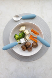 A white bowl with three meatballs, steamed broccoli, cauliflower, and carrot sticks sits on a gray placemat. On either side are a fork and spoon from the KiddiKutter Kids Cutlery Set [Sky Blue] by KiddiKutter.