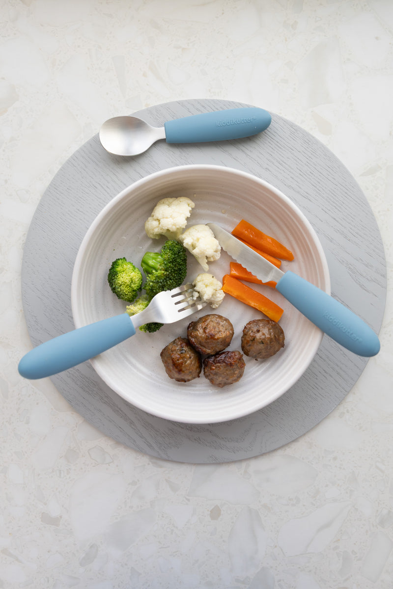 A white bowl with three meatballs, steamed broccoli, cauliflower, and carrot sticks sits on a gray placemat. On either side are a fork and spoon from the KiddiKutter Kids Cutlery Set [Sky Blue] by KiddiKutter.