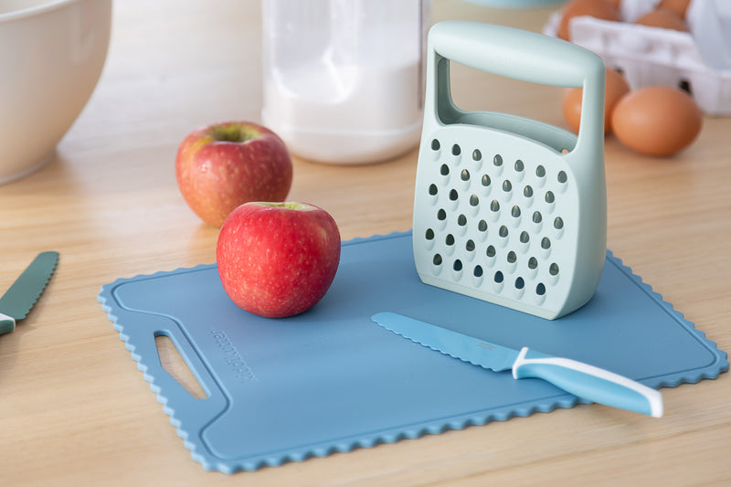 The KiddiKutter Silicone Cutting Board (Wavy) in Sky Blue by KiddiKutter, shown with apples, a blue-handled knife, grater, eggs in a carton, mixing bowl, and flour jar on wood—an ideal assistive product for household tasks.