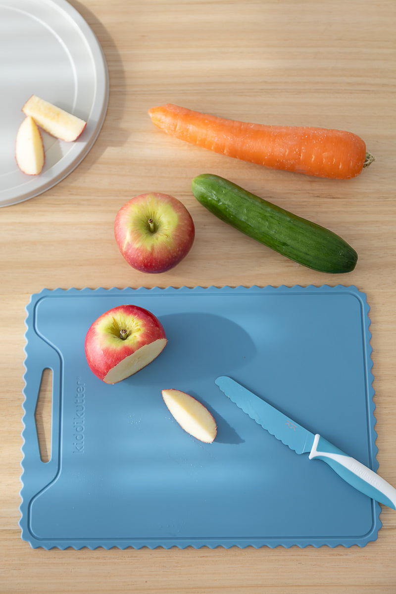 The KiddiKutter Silicone Cutting Board (Wavy) in Sky Blue by KiddiKutter sits on the table with a plastic knife, halved apple, apple slice, whole apple, cucumber, carrot, and a plate of more apple slices—perfect for household tasks.