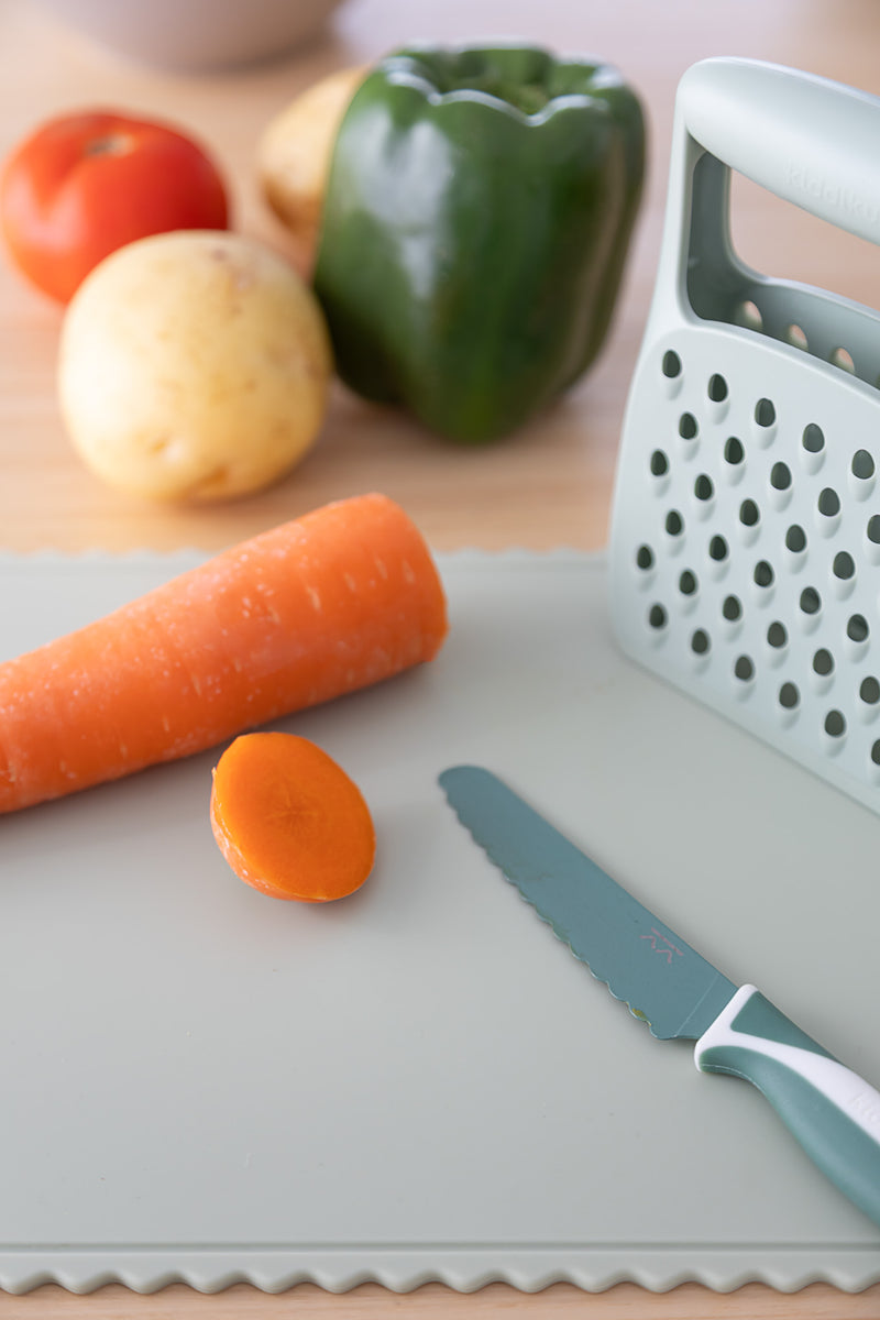 A sliced carrot, serrated knife, and box grater sit on the KiddiKutter Silicone Cutting Board (Wavy) [Sage]. A green bell pepper, tomato, and potato in the background make it perfect for fans of KiddiKutter's assistive kitchen products.