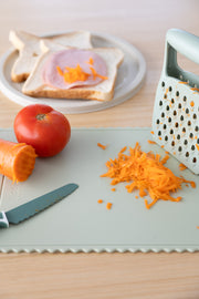 The KiddiKutter Silicone Cutting Board (Wavy) [Sage] by KiddiKutter is shown with a knife, grated carrot, shredded carrot, tomato, and grater. In the background, bread with ham and shredded carrot makes meal prep simple.