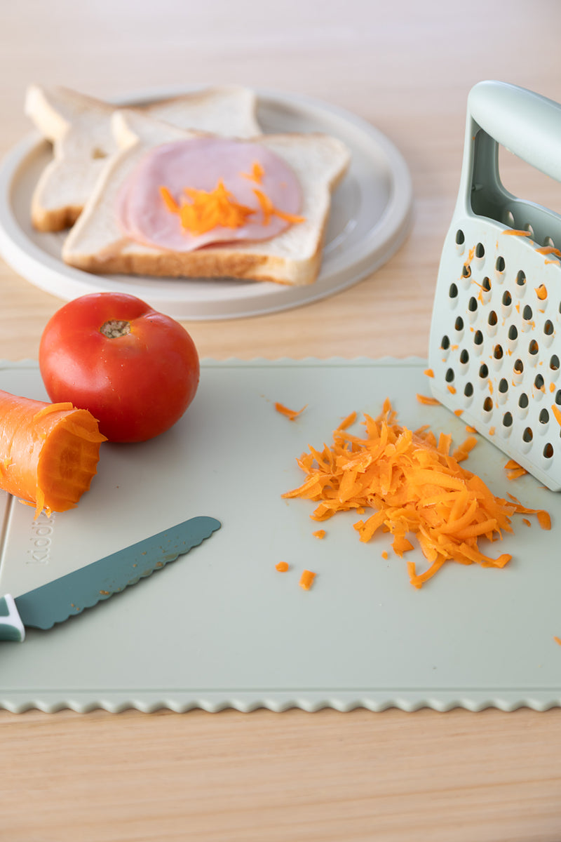 The KiddiKutter Silicone Cutting Board (Wavy) [Sage] by KiddiKutter is shown with a knife, grated carrot, shredded carrot, tomato, and grater. In the background, bread with ham and shredded carrot makes meal prep simple.
