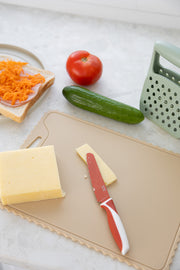 The KiddiKutter Silicone Cutting Board (Wavy) [Cream] by KiddiKutter holds cheese, a knife, grater, cucumber, tomato, and a plate with shredded cheese on toast—all neatly arranged on a light kitchen counter.