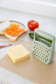 A block of cheese and a light green grater rest on the KiddiKutter Silicone Cutting Board (Wavy) in Cream. In the background, grated cheese, bread slices, a tomato, and a cucumber are visible.