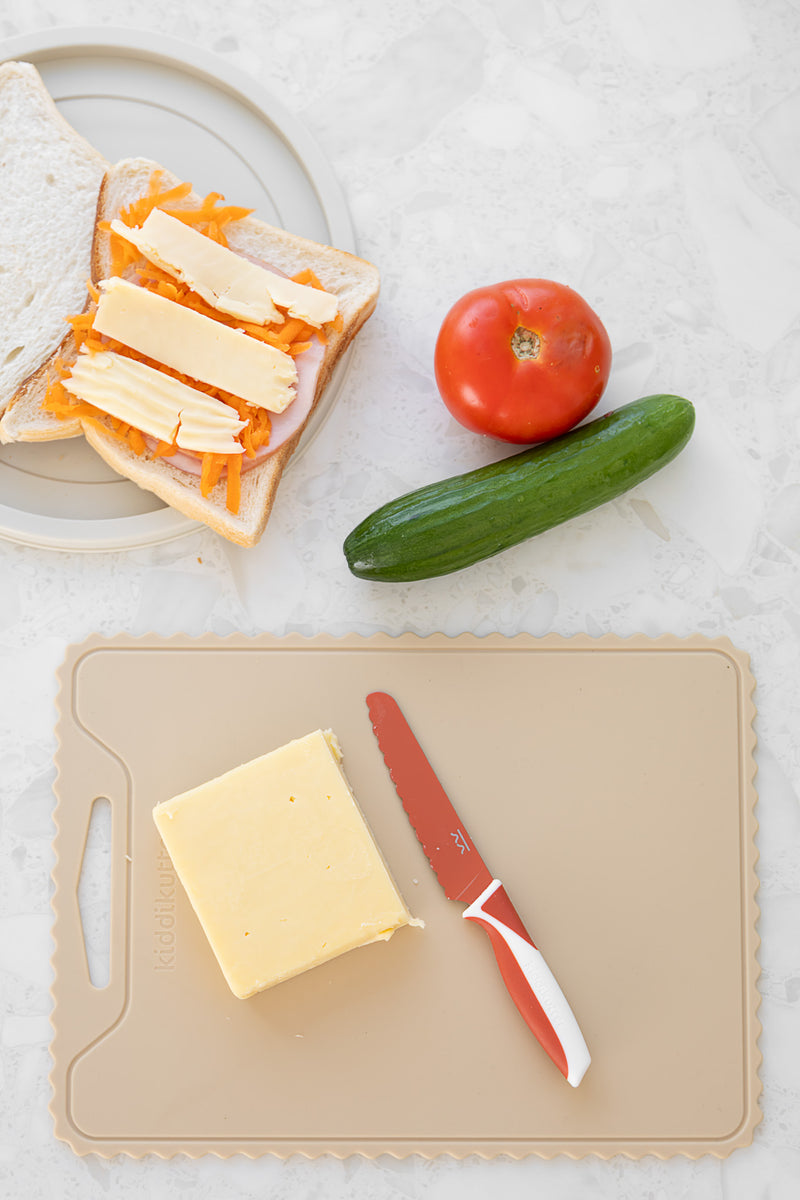 An open-faced sandwich with shredded carrots and cheese sits on a plate. Nearby, a tomato, cucumber, cheese block, and a red-handled knife rest on the KiddiKutter Silicone Cutting Board (Wavy) [Cream] by KiddiKutter on a light countertop.