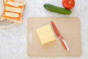 The KiddiKutter Silicone Cutting Board (Wavy) [Cream] by KiddiKutter securely holds cheese and a knife, with room for veggies and bread, making safe, easy food prep possible—perfect for assistive kitchen use.