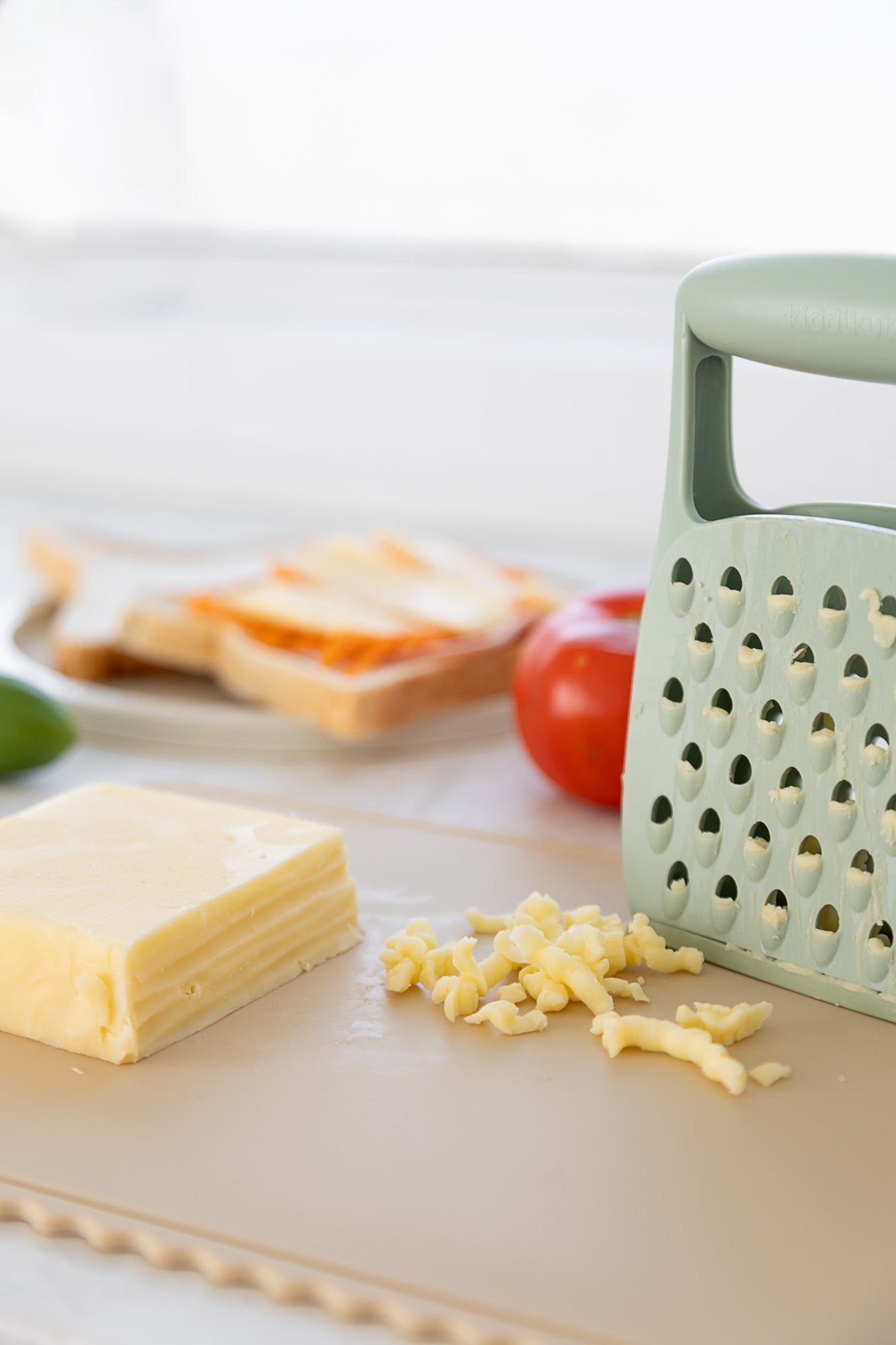 A block of cheese, shredded pieces, and a green KiddiKutter Grater by KiddiKutter sit on a cutting board. In the background, there are slices of bread, a tomato, and a green pepper slightly out of focus.