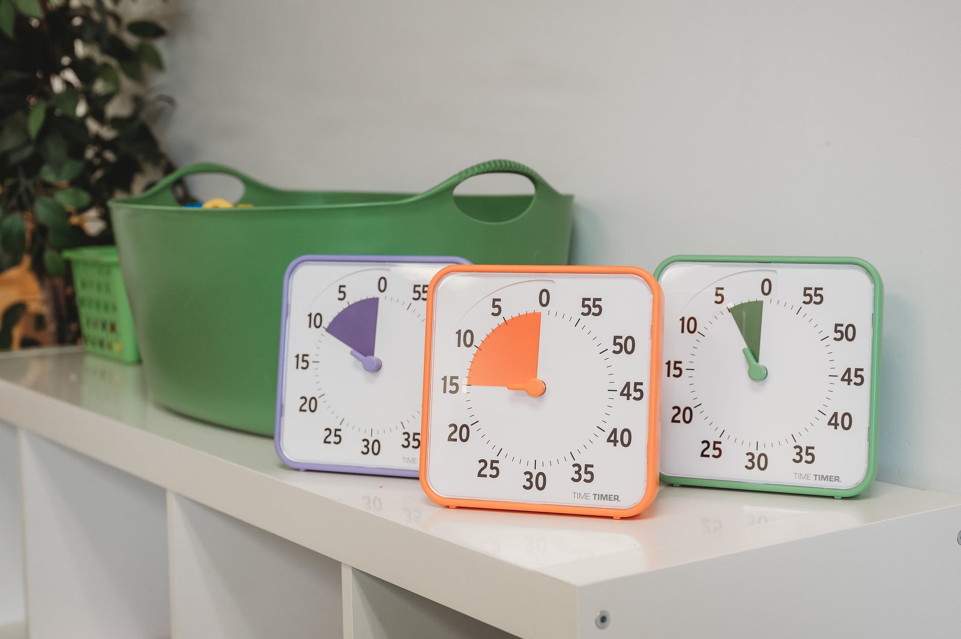 Three Time Timer Original (8 inch) Learning Center timers by Time Timer, from the Secondary Colour Collection, sit on a white shelf with a green storage bin and basket—perfect for classroom time management. A leafy plant appears in the background.