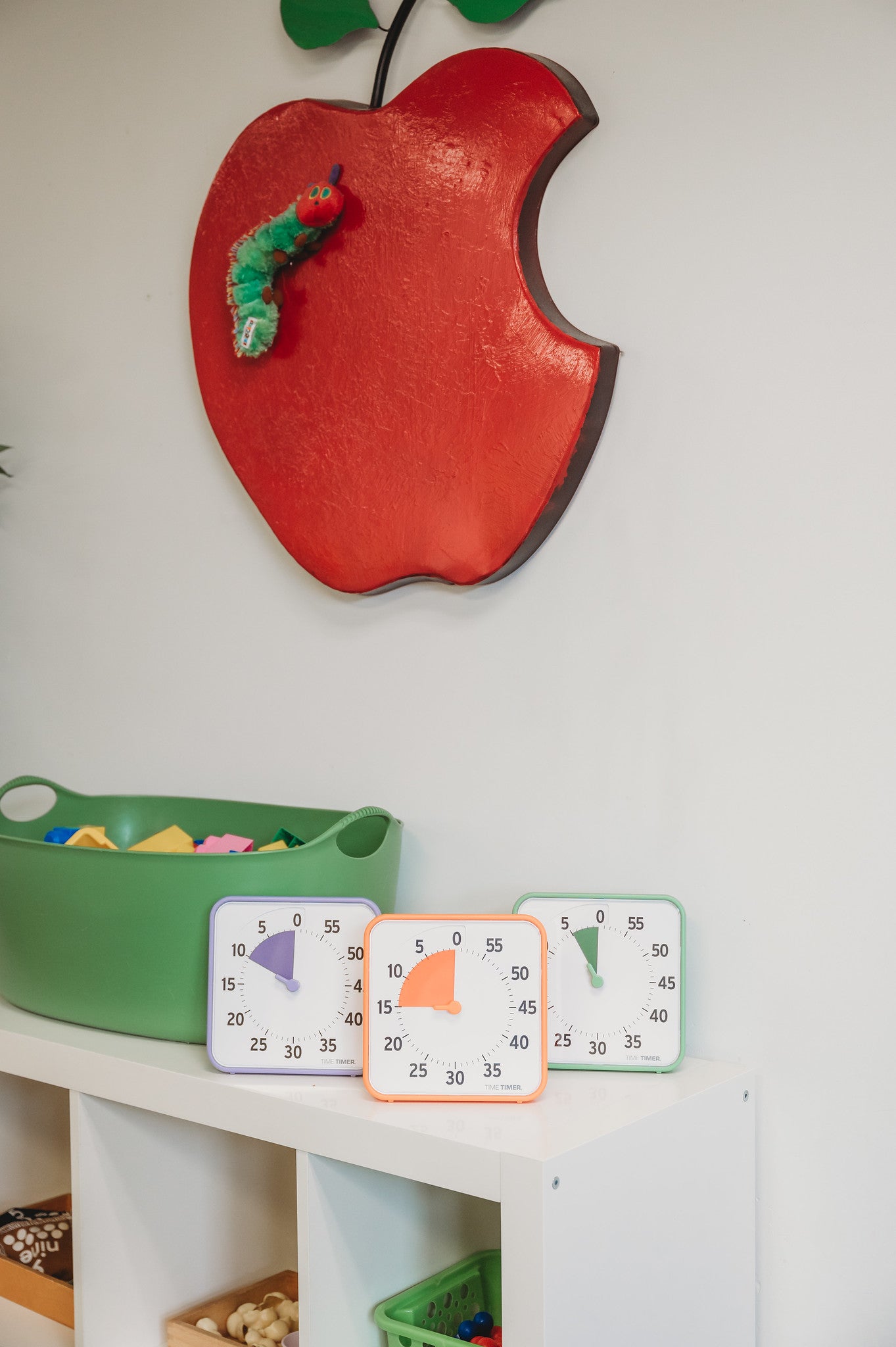 A classroom features a red apple wall decoration, a green basket with papers, and three Time Timer Original (8 inch) Learning Center Classroom Set timers by Time Timer—each with a different colored face for effective time management.