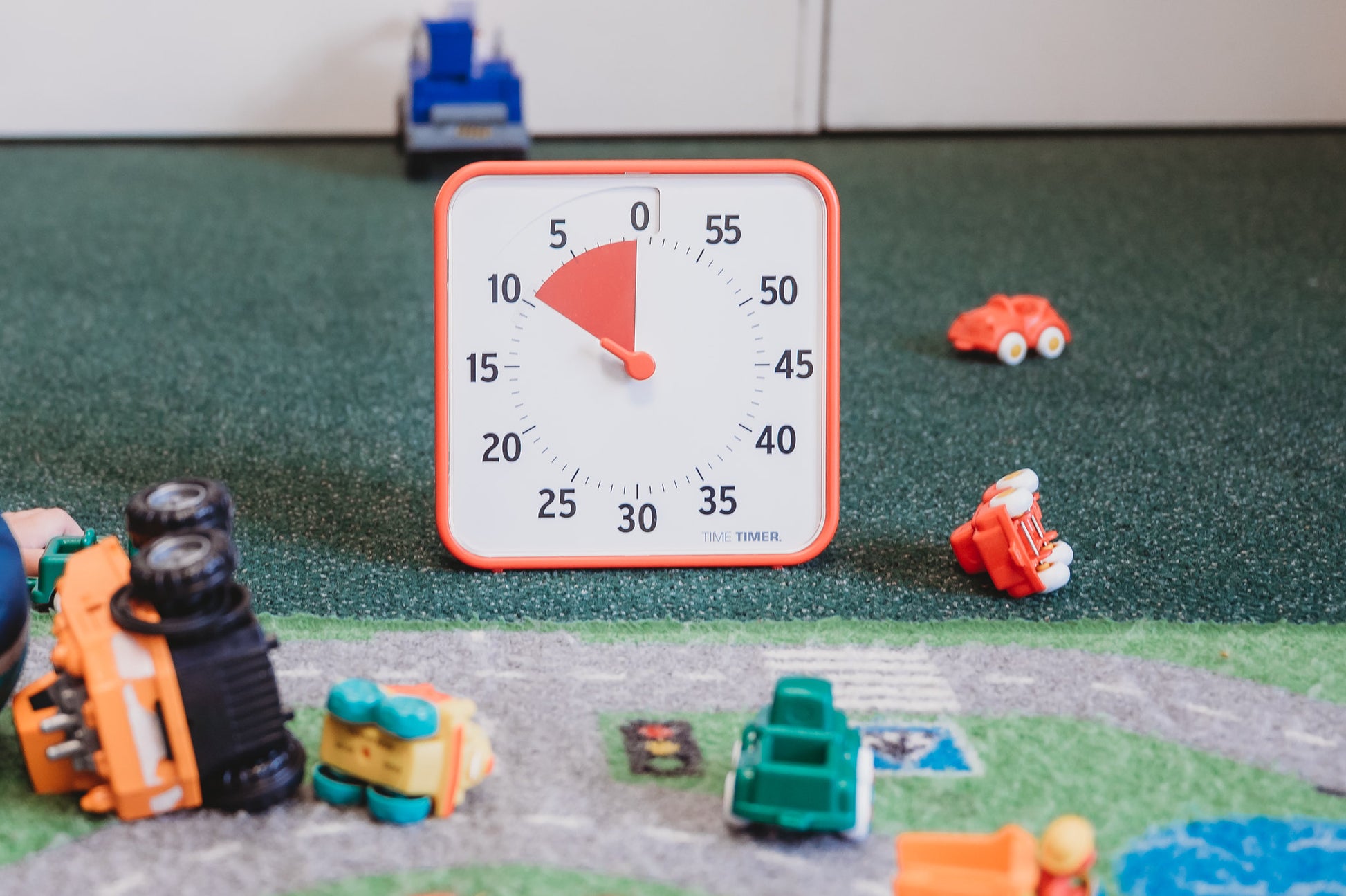 The Time Timer Original (8 inch) Learning Center Classroom Set (Secondary Colour Collection) by Time Timer, set to 5 minutes, sits on green carpet among toy cars and trucks. A toy road mat is partially visible in the foreground.